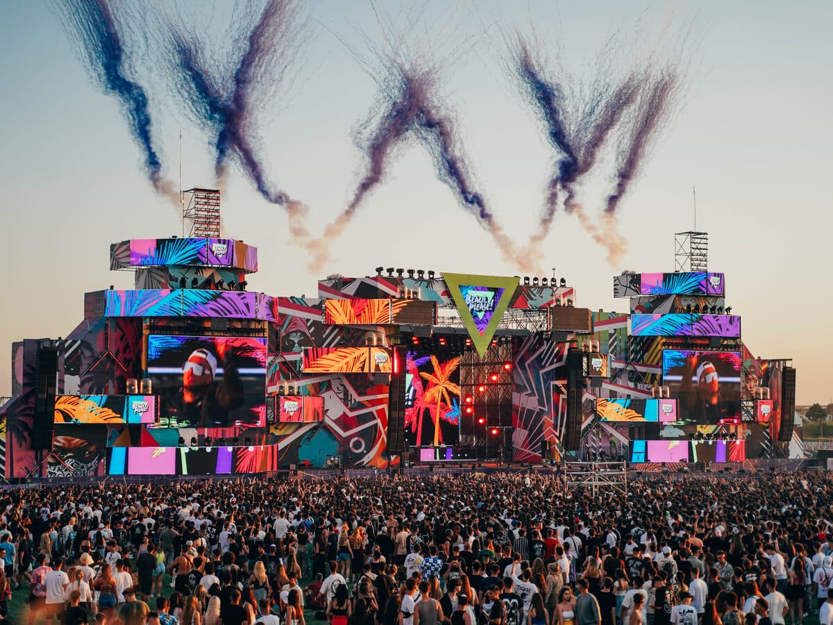 A crowd gathered in front of a large, colorful festival main stage at sunset