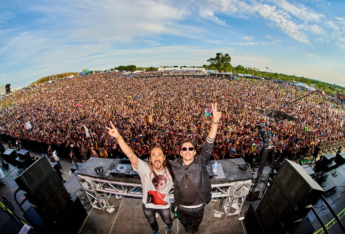 A DJ duo throwing peace signs to a massive daytime festival crowd